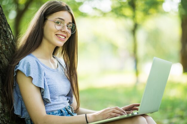 Mujer hermosa joven está sentada sobre la hierba verde debajo del árbol en el jardín el día de verano y trabajando en su computadora portátil