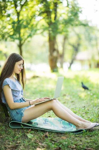 Mujer hermosa joven está sentada sobre la hierba verde debajo del árbol en el jardín el día de verano y trabajando en su computadora portátil