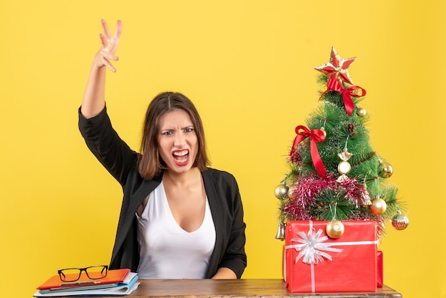 Mujer hermosa joven explicando algo emocionalmente sentado en una mesa cerca del árbol de Navidad decorado en la oficina en amarillo