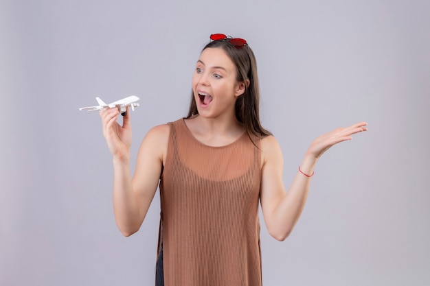 Mujer hermosa joven con gafas de sol rojas en la cabeza sosteniendo un avión de juguete mirando juguetón y feliz de pie con el brazo levantado sobre fondo blanco.