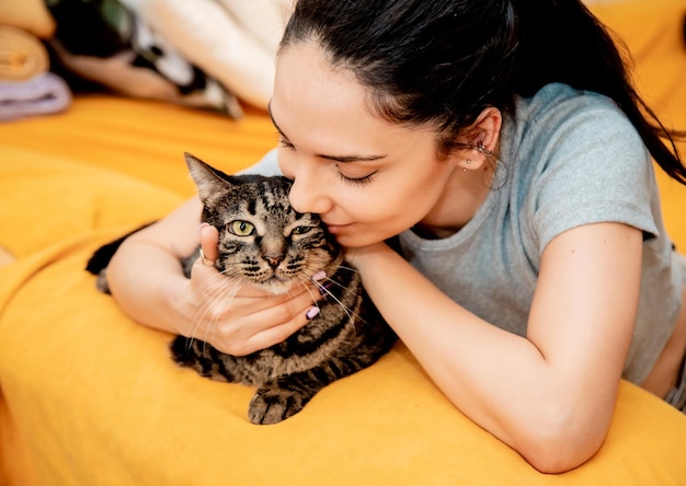 Mujer hermosa joven con gato en casa sentada en el sofá naranja abrazando a su linda mascota adorable concepto de mascota doméstica