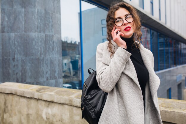 Mujer hermosa joven hablando por teléfono inteligente, estilo de la ciudad de la calle de otoño, abrigo, gafas, feliz, sonriente, sosteniendo el teléfono en la mano, cabello rizado