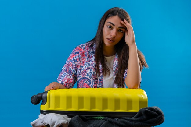 Mujer hermosa joven con una maleta llena de ropa que tiene dudas y con expresión de la cara confusa sobre la pared azul aislada