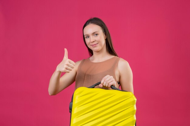 Mujer hermosa joven con maleta de viaje mirando a la cámara con cara feliz sonriendo mostrando los pulgares para arriba de pie sobre fondo rosa