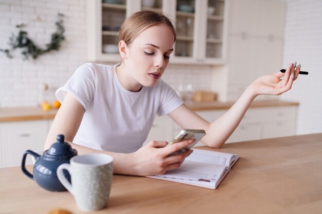 Mujer hermosa joven en la mañana en la cocina con un teléfono y un cuaderno