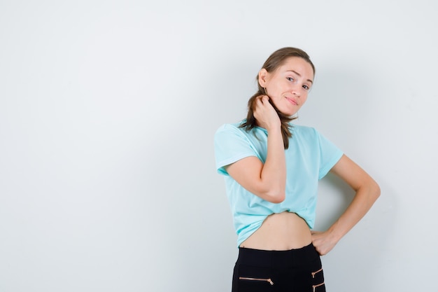 Mujer hermosa joven con la mano en la cintura, manteniendo la mano en la cintura en camiseta, pantalones y mirando feliz. vista frontal.