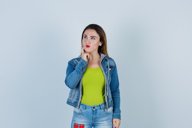 Mujer hermosa joven manteniendo el dedo en la mejilla, mirando a otro lado en traje de mezclilla y mirando pensativo, vista frontal.