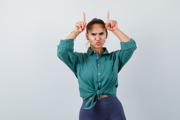 Mujer hermosa joven manteniendo las manos sobre la cabeza como cuernos con labios fruncidos en camisa verde y mirando pensativo. vista frontal.