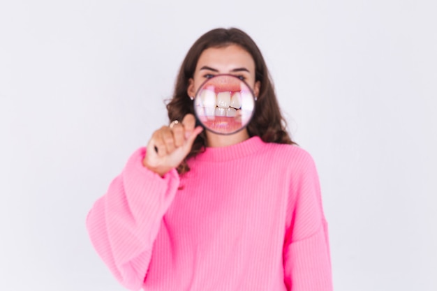 Mujer hermosa joven con maquillaje ligero pecas en suéter en la pared blanca con lupa muestra una sonrisa perfecta de dientes blancos