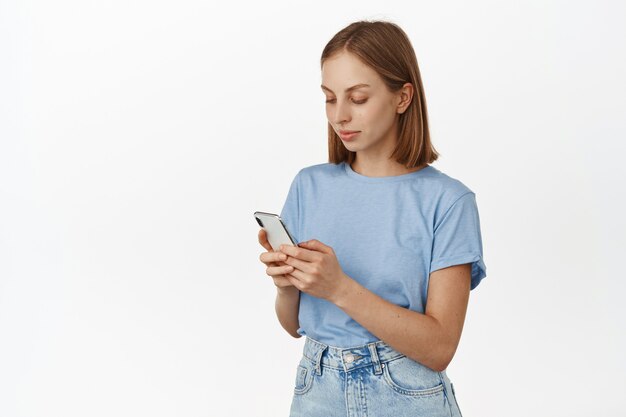 Mujer hermosa joven con el mensaje de la escritura del pelo corto en el teléfono móvil. Mujer mirando teléfono inteligente, enviando mensajes de texto o chateando, aplicación de redes sociales de desplazamiento, pared blanca.