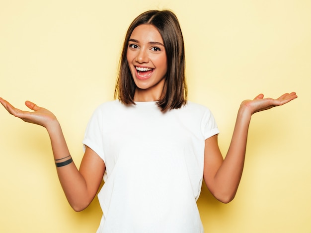 Mujer hermosa joven mirando a cámara. Chica de moda en camiseta blanca de verano casual. La hembra positiva muestra emociones faciales. Modelo que muestra algo en ambas manos planas para una elección similar de producto