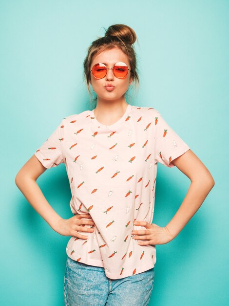 Mujer hermosa joven mirando a cámara. Chica de moda en ropa casual de camiseta de verano hace cara de pato. La hembra positiva muestra emociones faciales. Modelo divertido aislado en azul en gafas de sol