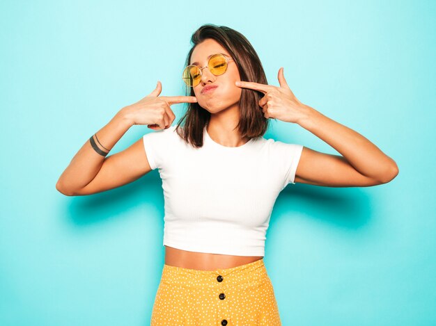 Mujer hermosa joven mirando a cámara. Chica de moda en verano casual camiseta blanca y falda amarilla en gafas de sol redondas. La hembra positiva muestra emociones faciales. Modelo divertido soplando sus mejillas.