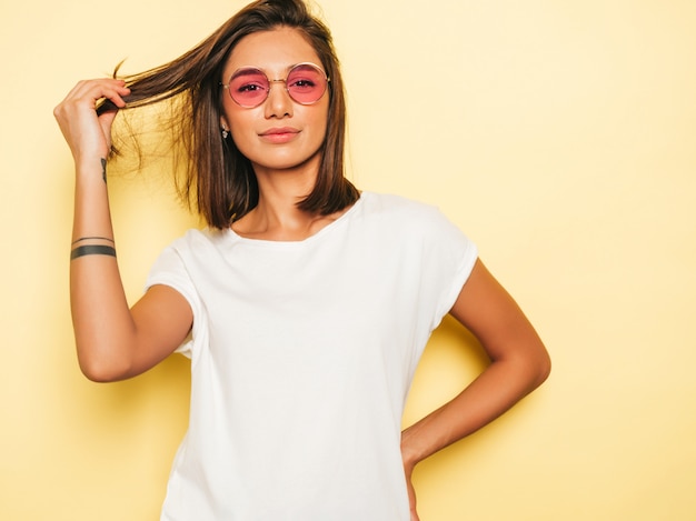 Mujer hermosa joven mirando a cámara. Chica de moda en verano casual camiseta blanca y pantalones vaqueros en gafas de sol redondas. La hembra positiva muestra emociones faciales. Modelo divertido aislado en amarillo