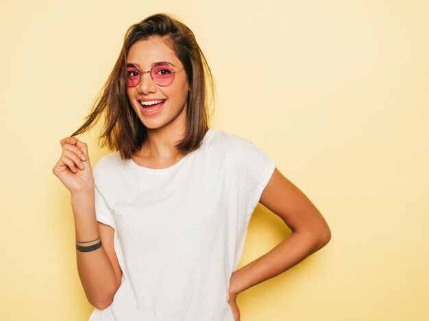 Mujer hermosa joven mirando a cámara. Chica de moda en verano casual camiseta blanca y pantalones vaqueros en gafas de sol redondas. La hembra positiva muestra emociones faciales. Modelo divertido aislado en amarillo