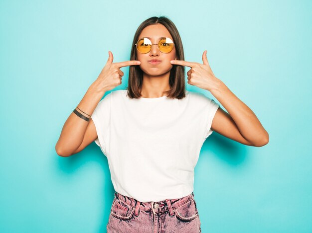 Mujer hermosa joven mirando a cámara. Chica de moda en verano casual camiseta blanca y pantalones vaqueros en gafas de sol redondas. La hembra positiva muestra emociones faciales. Modelo divertido soplando sus mejillas.