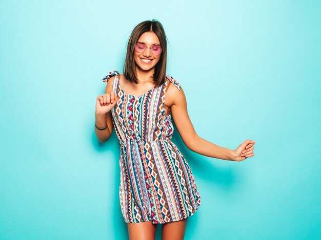 Mujer hermosa joven mirando a cámara. Chica de moda en vestido casual de verano y con gafas de sol redondas. La hembra positiva muestra emociones faciales. Modelo divertido aislado en azul