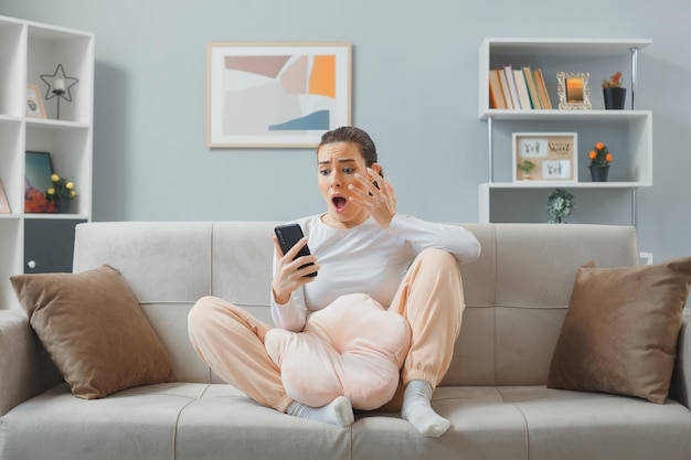 Mujer hermosa joven molesta con ropa informal sentada en un sofá en el interior de su casa sosteniendo un mensaje de lectura de teléfono inteligente sorprendida y disgustada levantando el brazo con disgusto