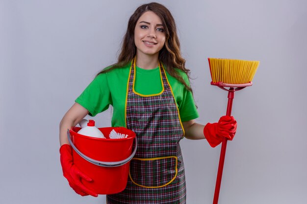 Mujer hermosa joven con pelo largo ondulado en delantal y guantes con balde con herramientas de limpieza y fregona sonriendo alegre de pie