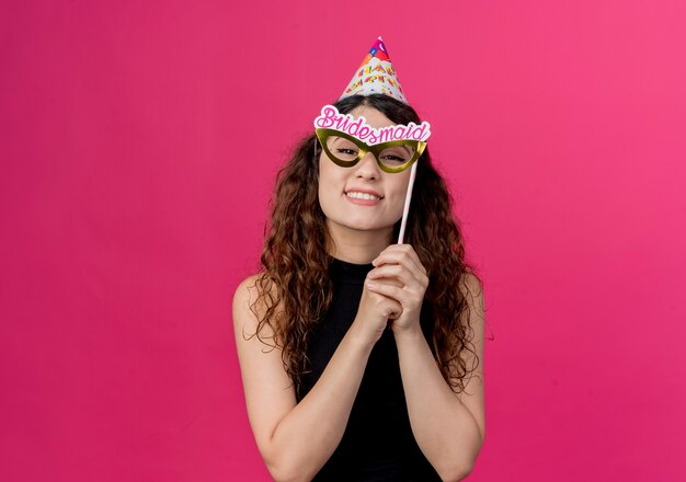 Mujer hermosa joven con el pelo rizado celebración palo de fiesta feliz y alegre sonriendo de pie sobre la pared rosa