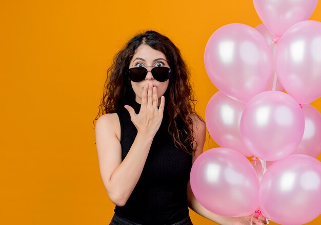 Mujer hermosa joven con el pelo rizado en gafas negras sosteniendo un montón de globos de aire sorprendidos y asombrados de pie sobre la pared naranja