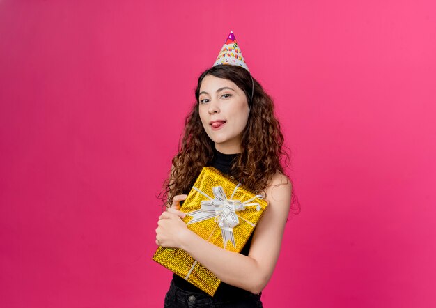 Mujer hermosa joven con el pelo rizado en una gorra de vacaciones con caja de regalo de cumpleaños sonriendo alegremente sacando la lengua concepto de fiesta de cumpleaños de pie sobre la pared rosa