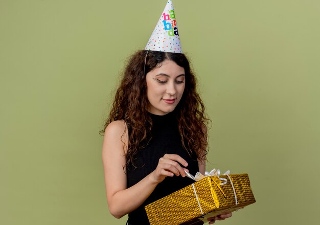 Mujer hermosa joven con el pelo rizado en una gorra de vacaciones con caja de regalo mirándola con una sonrisa en la cara concepto de fiesta de cumpleaños de pie sobre una pared ligera