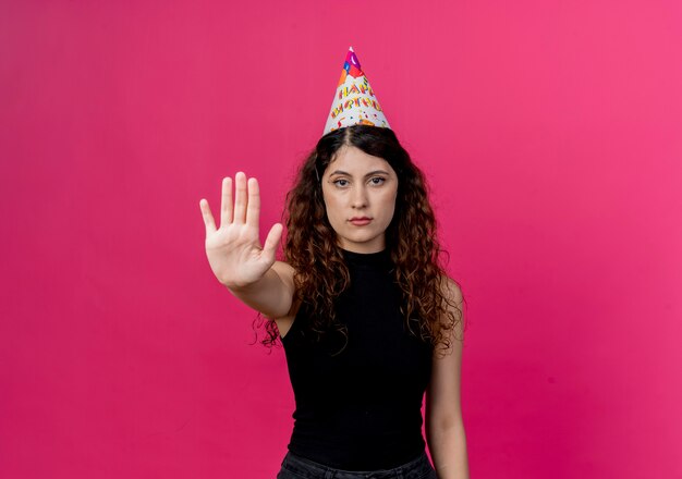 Mujer hermosa joven con el pelo rizado en una gorra de vacaciones con cara seria haciendo señal de stop concepto de fiesta de cumpleaños de pie sobre la pared rosa