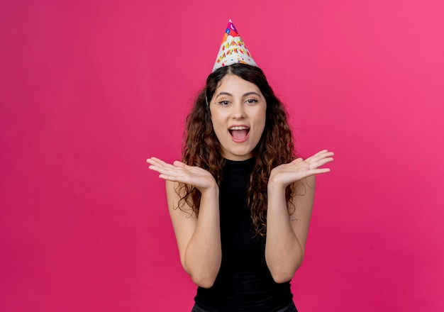 Mujer hermosa joven con el pelo rizado en una gorra de vacaciones concepto de fiesta de cumpleaños sorprendido y feliz parado sobre pared rosa