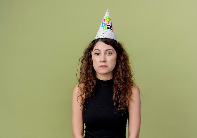 Mujer hermosa joven con el pelo rizado en una gorra de vacaciones con expresión triste en la cara concepto de fiesta de cumpleaños de pie sobre la pared naranja