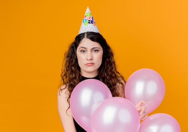 Mujer hermosa joven con el pelo rizado en una gorra de vacaciones con globos de aire concepto de fiesta de cumpleaños disgustado e infeliz de pie sobre la pared naranja
