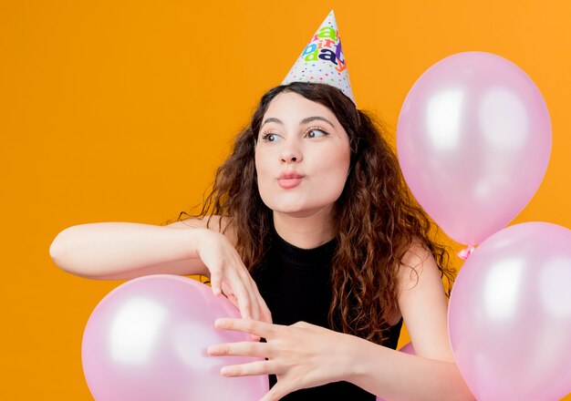 Mujer hermosa joven con el pelo rizado en una gorra de vacaciones con globos de aire concepto de fiesta de cumpleaños feliz y emocionado de pie sobre la pared naranja