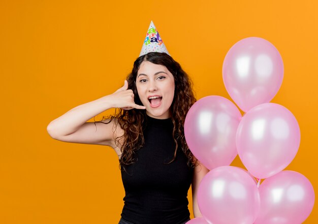 Mujer hermosa joven con el pelo rizado en una gorra de vacaciones con globos de aire mostrando feliz y positivo concepto de fiesta de cumpleaños de gesto de llamarme de pie sobre la pared naranja