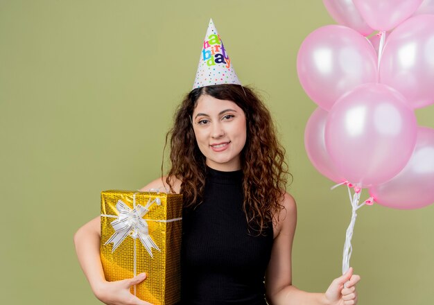 Mujer hermosa joven con el pelo rizado en una gorra de vacaciones con globos de aire y regalo de cumpleaños sonriendo con cara feliz de pie sobre la pared de luz