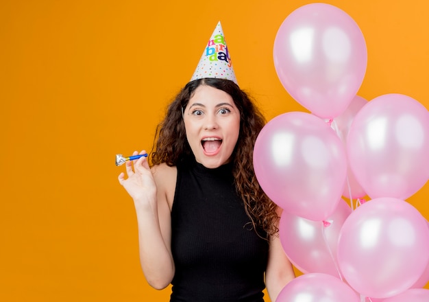 Mujer hermosa joven con el pelo rizado en una gorra de vacaciones con globos de aire y silbido feliz y emocionado sonriendo ampliamente concepto de fiesta de cumpleaños de pie sobre la pared naranja