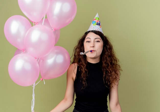 Mujer hermosa joven con el pelo rizado en una gorra de vacaciones con globos de aire soplando silbato feliz y positiva celebración de cumpleaños de pie sobre pared ligera