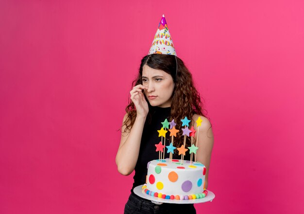 Mujer hermosa joven con el pelo rizado en una gorra de vacaciones con pastel de cumpleaños mirando a un lado confundido concepto de fiesta de cumpleaños de pie sobre la pared rosa