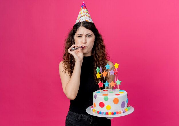 Mujer hermosa joven con el pelo rizado en una gorra de vacaciones con pastel de cumpleaños que sopla silbato concepto de fiesta de cumpleaños feliz y positivo de pie sobre la pared rosa