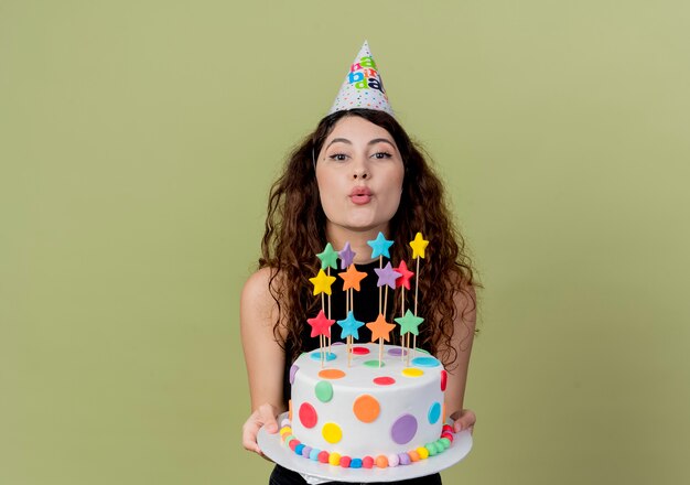 Mujer hermosa joven con el pelo rizado en una gorra de vacaciones con pastel de cumpleaños sonriendo alegremente feliz y alegre sobre la luz