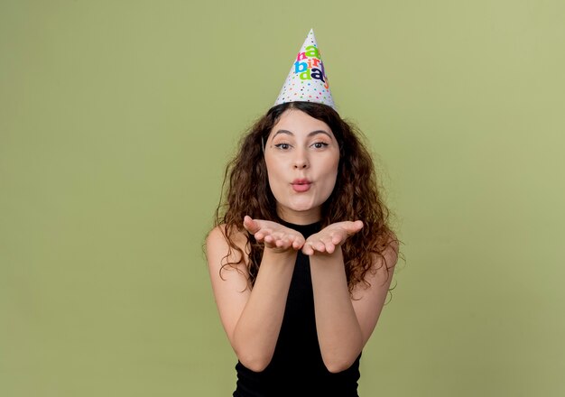Mujer hermosa joven con el pelo rizado en una gorra de vacaciones que sopla un beso con las manos delante de su concepto de fiesta de cumpleaños de pie sobre una pared de luz