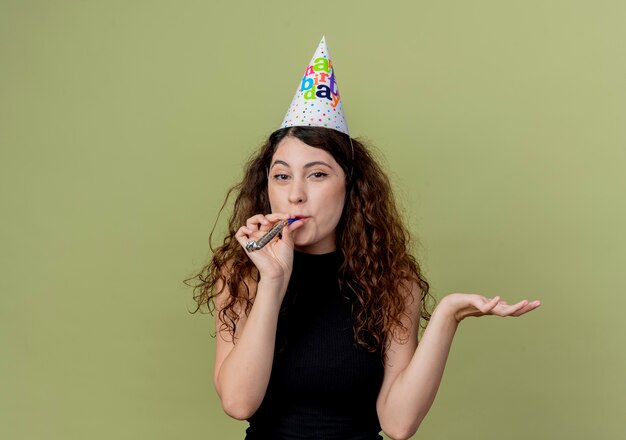 Mujer hermosa joven con el pelo rizado en una gorra de vacaciones que sopla el silbato con el brazo extendido concepto de fiesta de cumpleaños de pie sobre una pared de luz