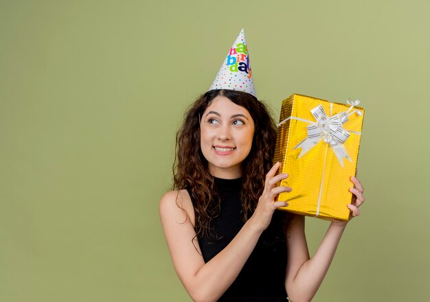 Foto gratuita mujer hermosa joven con el pelo rizado en una gorra de vacaciones con regalo de cumpleaños concepto de fiesta de cumpleaños feliz y positivo de pie sobre la pared de luz