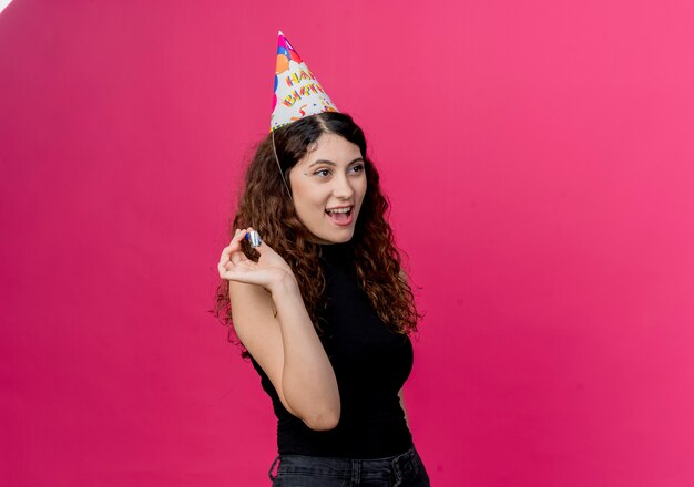 Mujer hermosa joven con el pelo rizado en una gorra de vacaciones con silbato concepto de fiesta de cumpleaños feliz y positivo de pie sobre la pared rosa