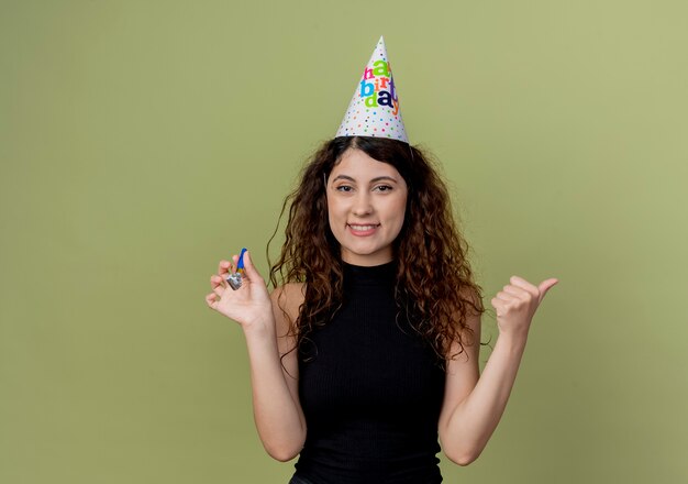 Mujer hermosa joven con el pelo rizado en una gorra de vacaciones con silbato sonriendo alegremente concepto de fiesta de cumpleaños de pie sobre la pared de luz