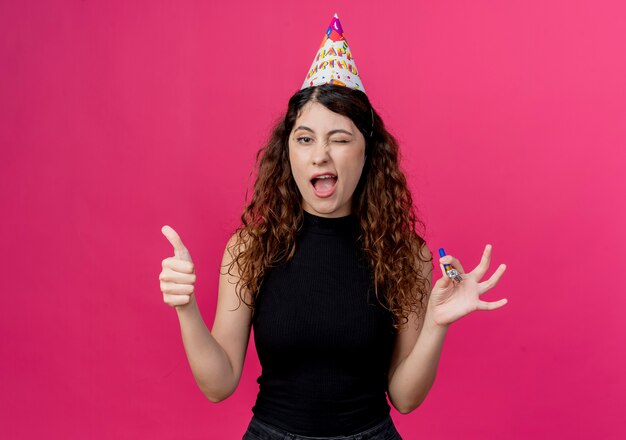 Mujer hermosa joven con el pelo rizado en una gorra de vacaciones sonriendo y guiñando un ojo mostrando los pulgares para arriba concepto de fiesta de cumpleaños de pie sobre la pared rosa