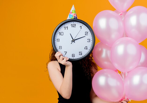 Mujer hermosa joven con el pelo rizado en una gorra de vacaciones sosteniendo globos de aire escondiendo la cara detrás del reloj de pared concepto de fiesta de cumpleaños de pie sobre la pared naranja