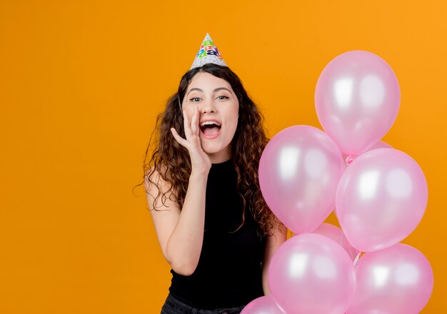 Mujer hermosa joven con el pelo rizado en una gorra de vacaciones sosteniendo globos de aire gritando o llamando con la mano cerca de la boca concepto de fiesta de cumpleaños sobre naranja
