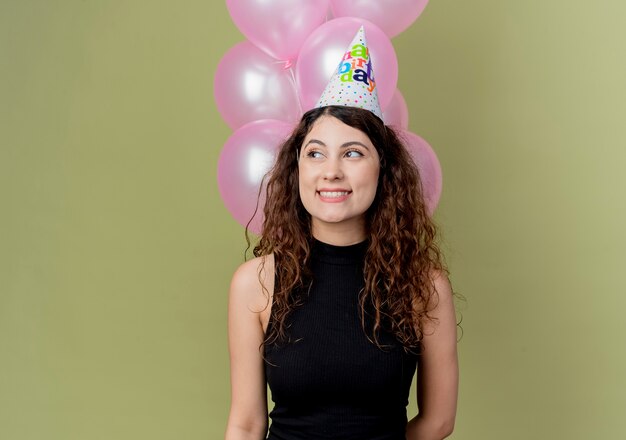 Mujer hermosa joven con el pelo rizado en una gorra de vacaciones sosteniendo globos de aire mirando a un lado sonriendo alegremente con cara feliz celebrando la fiesta de cumpleaños de pie sobre la pared de luz