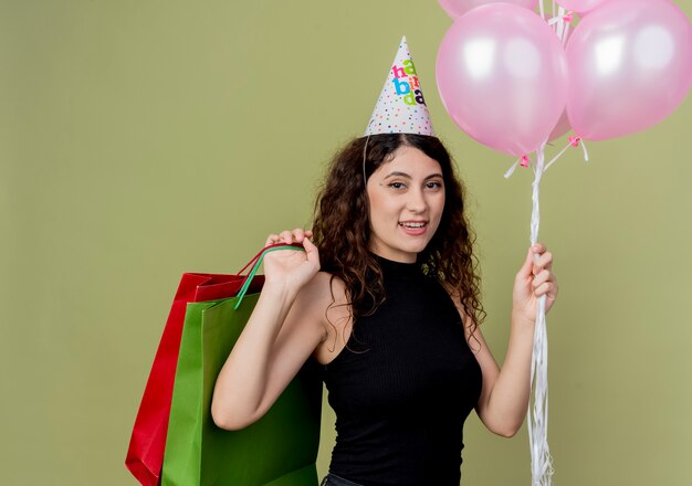 Mujer hermosa joven con el pelo rizado en una gorra de vacaciones sosteniendo globos de aire y regalos de cumpleaños feliz y positivo sonriendo alegremente de pie sobre la pared de luz