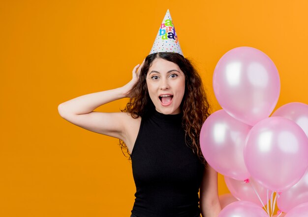 Mujer hermosa joven con el pelo rizado en una gorra de vacaciones sosteniendo globos de aire sorprendido y feliz cumpleaños concepto de fiesta de pie sobre la pared naranja